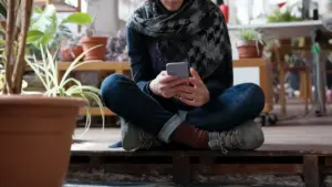 Una persona seduta a terra guarda lo smartphone in un ambiente domestico con piante