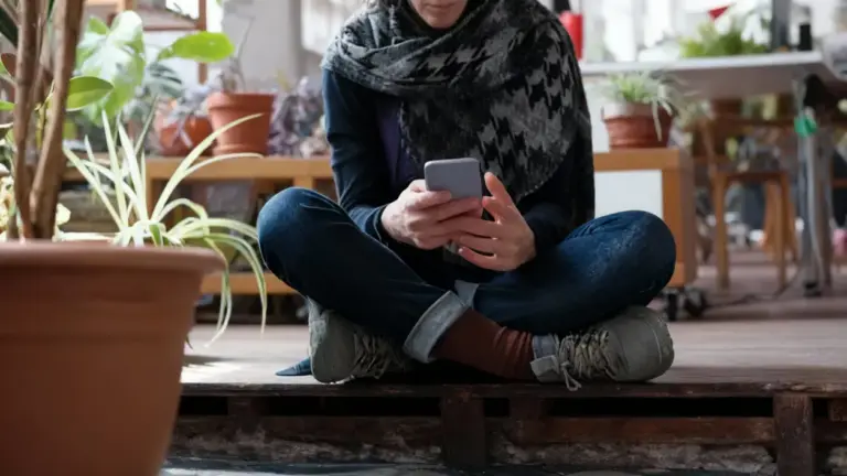 Una persona seduta a terra guarda lo smartphone in un ambiente domestico con piante